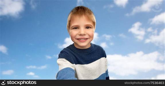 childhood, expressions and people concept - portrait of nice little boy in striped pullover taking selfie over blue sky and clouds background. happy little boy in striped pullover taking selfie