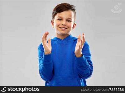 childhood, expressions and people concept - portrait of smiling little boy in blue hoodie applauding over grey background. portrait of smiling boy in blue hoodie applauding