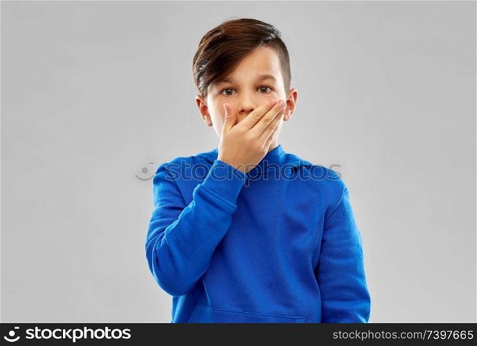 childhood, expressions and people concept - shocked little boy in blue hoodie closing his mouth by hand over grey background. shocked boy closing mouth by hand