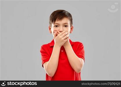 childhood, expressions and people concept - shocked little boy in red polo t-shirt closing his mouth by hands over grey background. shocked boy in red t-shirt closing mouth by hands