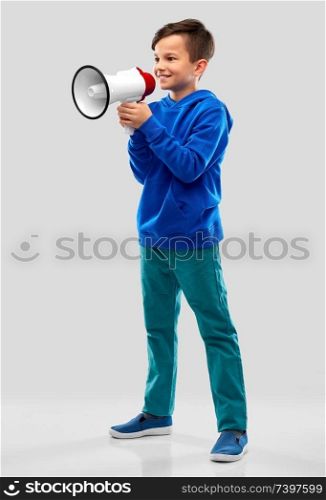 childhood, expressions and people concept - smiling boy in blue hoodie speaking to megaphone over grey background. smiling boy speaking to megaphone