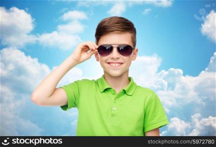 childhood, fashion, accessory, style and people concept - happy smiling boy in sunglasses and green polo t-shirt over blue sky and clouds background