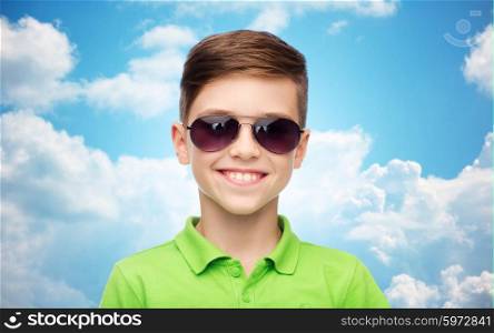 childhood, fashion, accessory, style and people concept - happy smiling boy in sunglasses and green polo t-shirt over blue sky and clouds background