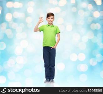 childhood, fashion, advertisement and people concept - happy boy in white t-shirt and jeans showing ok hand sign over blue holidays lights background