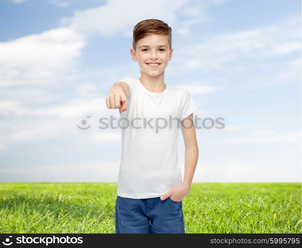 childhood, fashion, advertisement and people concept - happy boy in white t-shirt and jeans pointing finger to you over blue sky and grass background