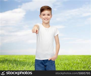 childhood, fashion, advertisement and people concept - happy boy in white t-shirt and jeans pointing finger to you over blue sky and grass background