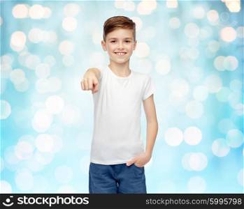 childhood, fashion, advertisement and people concept - happy boy in white t-shirt and jeans pointing finger to you over blue holidays lights background