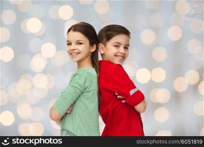 childhood, fashion and people concept - happy smiling boy and girl standing back to back over holidays lights background