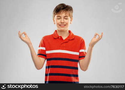 childhood, fashion and people concept - happy smiling boy in red polo t-shirt meditating over grey background. happy smiling meditating boy in red polo t-shirt
