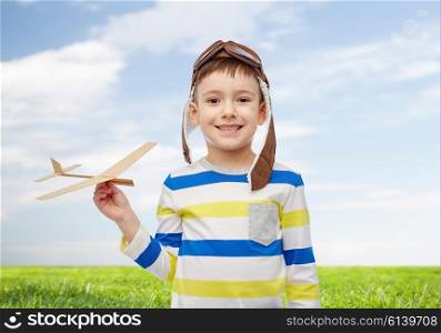 childhood, fashion and people concept - happy smiling little boy in aviator hat playing with wooden airplane over blue sky and green field background