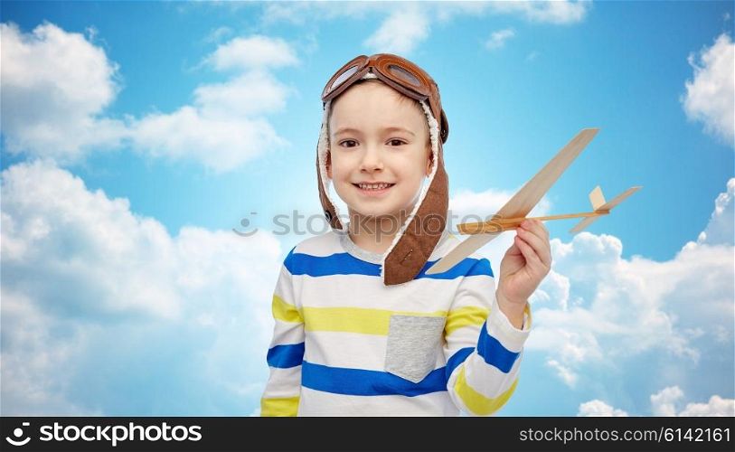 childhood, fashion and people concept - happy smiling little boy in aviator hat playing with wooden airplane over blue sky and clouds background