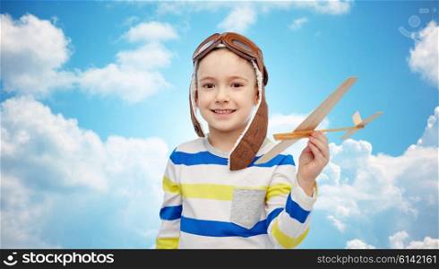 childhood, fashion and people concept - happy smiling little boy in aviator hat playing with wooden airplane over blue sky and clouds background