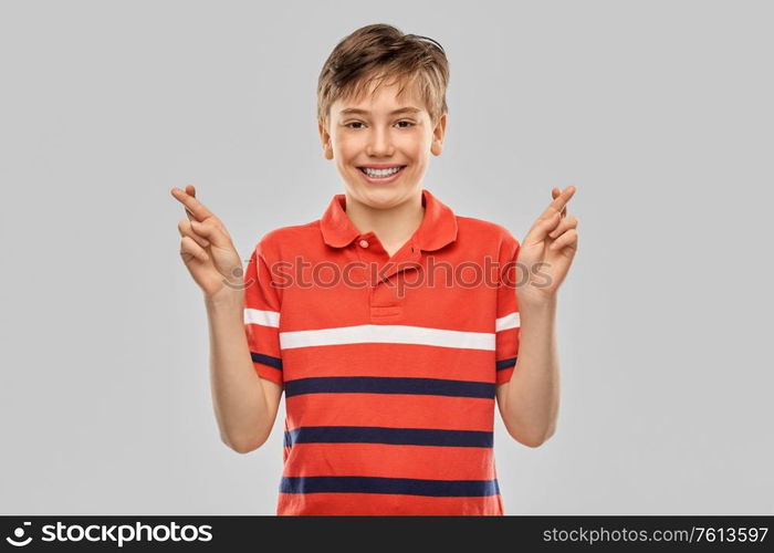 childhood, fashion and people concept - portrait of happy smiling boy in red polo t-shirt holding fingers crossed over grey background. smiling boy in t-shirt holding fingers crossed