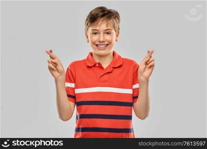 childhood, fashion and people concept - portrait of happy smiling boy in red polo t-shirt holding fingers crossed over grey background. smiling boy in t-shirt holding fingers crossed