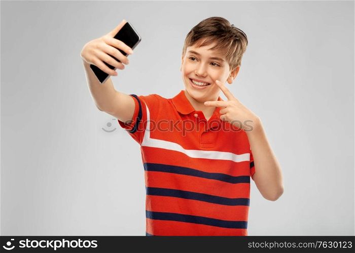 childhood, fashion and people concept - portrait of happy smiling boy in red polo t-shirt taking selfie with smartphone and showing peace over grey background. happy smiling boy taking selfie with smartphone