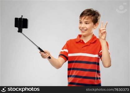 childhood, fashion and people concept - portrait of happy smiling boy in red polo t-shirt taking picture with smartphone on selfie stick and showing peace over grey background. boy taking picture with smartphone on selfie stick