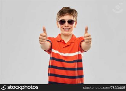 childhood, fashion and people concept - portrait of happy smiling boy in sunglasses and red polo t-shirt showing thumbs up over grey background. smiling boy in sunglasses showing thumbs up