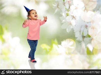 childhood, fashion, birthday, holidays and people concept - happy smiling african american little baby girl with birthday party hat playing and catching something over cherry blossoms background