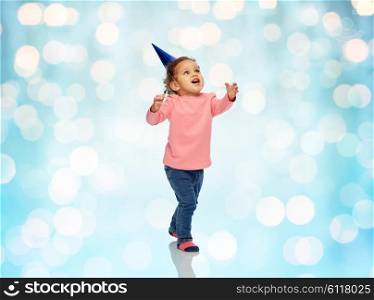 childhood, fashion, birthday, holidays and people concept - happy smiling african american little baby girl with birthday party hat playing and catching something over blue holidays lights background