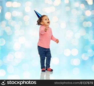 childhood, fashion, birthday, holidays and people concept - happy smiling african american little baby girl with birthday party hat looking up at something over blue holidays lights background