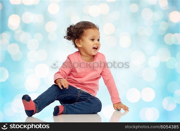 childhood, fashion, clothing and people concept - smiling beautiful african american little baby girl sitting on floor over blue holidays lights background