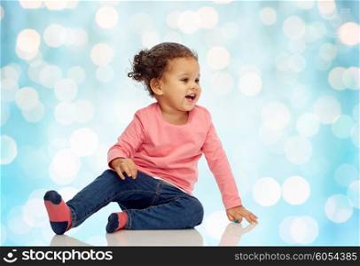 childhood, fashion, clothing and people concept - smiling beautiful african american little baby girl sitting on floor over blue holidays lights background