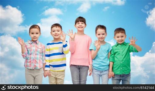 childhood, fashion, friendship and people concept - group of happy smiling little children holding hands over blue sky and clouds background