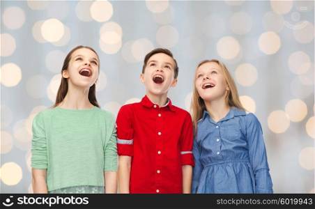 childhood, fashion, friendship and people concept - happy amazed boy and girls looking up with open mouths over holidays lights background