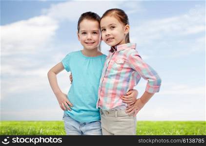 childhood, fashion, friendship and people concept - happy smiling little girls hugging over blue sky and green field background
