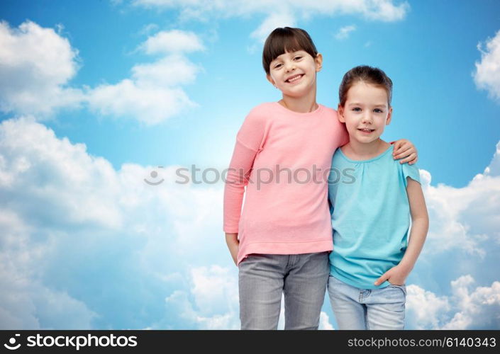 childhood, fashion, friendship and people concept - happy smiling little girls hugging over blue sky and clouds background