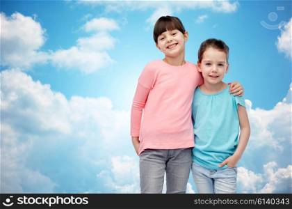 childhood, fashion, friendship and people concept - happy smiling little girls hugging over blue sky and clouds background