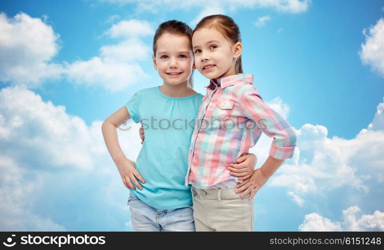 childhood, fashion, friendship and people concept - happy smiling little girls hugging over blue sky and clouds background