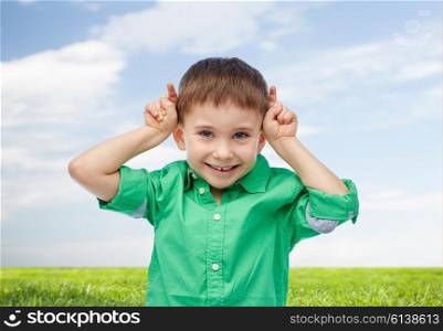 childhood, fashion, fun and people concept - happy little boy having fun and making horns over blue sky and green field background