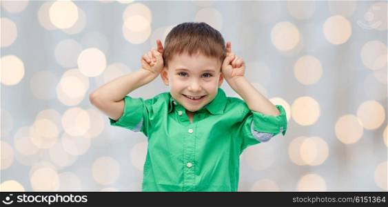 childhood, fashion, fun and people concept - happy little boy having fun and making horns over holidays lights background