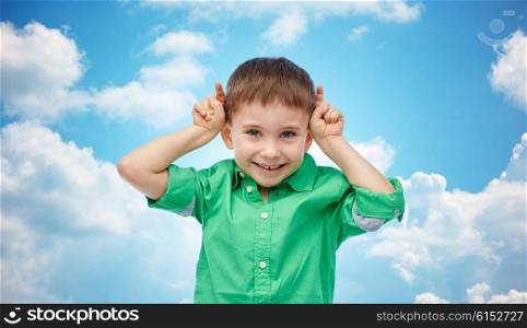 childhood, fashion, fun and people concept - happy little boy having fun and making horns over blue sky and clouds background