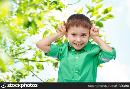 childhood, fashion, fun, summer and people concept - happy little boy having fun and making horns over green natural background