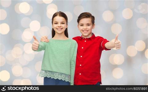 childhood, fashion, gesture and people concept - happy smiling boy and girl hugging and showing thumbs up over holidays lights background