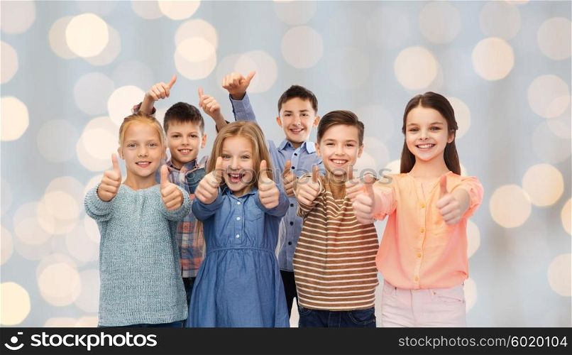 childhood, fashion, gesture and people concept - happy smiling children showing thumbs up over holidays lights background