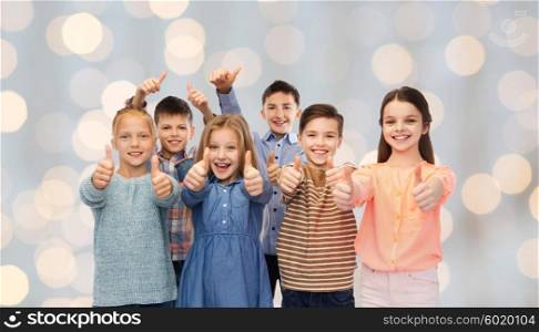 childhood, fashion, gesture and people concept - happy smiling children showing thumbs up over holidays lights background