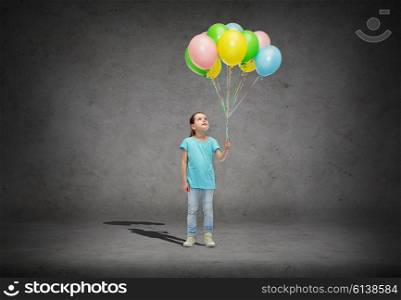 childhood, fashion, imagination and people concept - happy little girl looking up and holding bunch of colorful helium balloons on strand over concrete room background