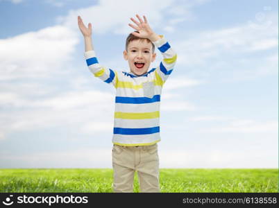 childhood, fashion, joy and people concept - happy little boy waving hands over blue sky and green field background