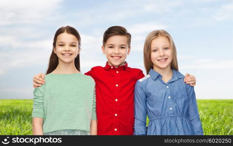 childhood, fashion, summer, friendship and people concept - happy smiling boy and girls hugging over blue sky and grass background