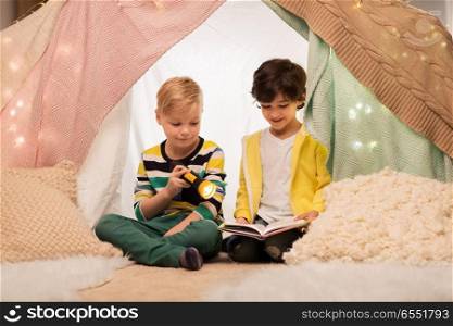 childhood, friendship and hygge concept - happy little boys reading book with torch light in kids tent or teepee at home. happy boys reading book in kids tent at home. happy boys reading book in kids tent at home