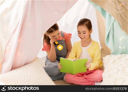 childhood, friendship and hygge concept - happy little girls reading book with torch light in kids tent or teepee at home. little girls reading book in kids tent at home. little girls reading book in kids tent at home