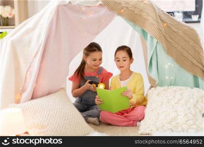 childhood, friendship and hygge concept - happy little girls reading book with torch light in kids tent or teepee at home. little girls reading book in kids tent at home