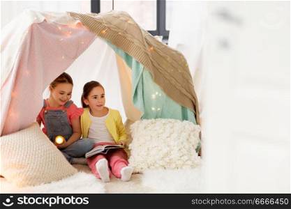 childhood, friendship and hygge concept - happy little girls with book and torch light in kids tent or teepee at home. girls with book and torch in kids tent at home. girls with book and torch in kids tent at home