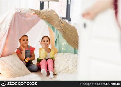 childhood, friendship and hygge concept - happy little girls with book and torch light waving hands to parent in kids tent or teepee at home. girls with book and torch in kids tent at home. girls with book and torch in kids tent at home