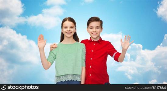 childhood, friendship, gesture and people concept - happy smiling boy and girl hugging and waving hand over blue sky and clouds background