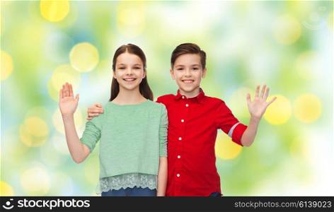 childhood, friendship, gesture and people concept - happy smiling boy and girl hugging and waving hand over green summer holidays lights background