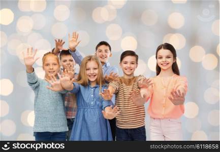 childhood, friendship, gesture and people concept - happy smiling children waving hands over holidays lights background